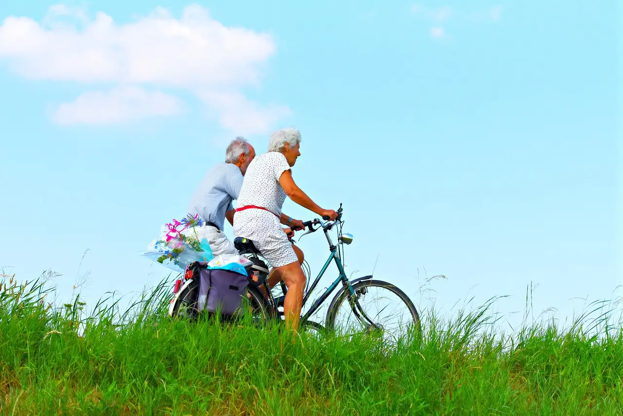 Two older people happily ride their bikes uphill through bright green grass under a clear blue sky. They look full of energy and health. One bike has a basket with flowers in it, and the shadows from the flowers stand out in the sunny day, showing a calm and active scene. Subscribe For a Free eBook